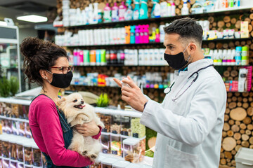 Female customer with protective face mask talking with veterinarian in pet shop and holding cute Pomeranian dog.