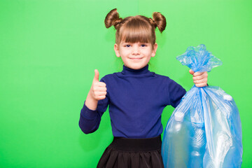 Happy little girl holding garbage bag with plastic trash and showing winning gesture isolated on green background
