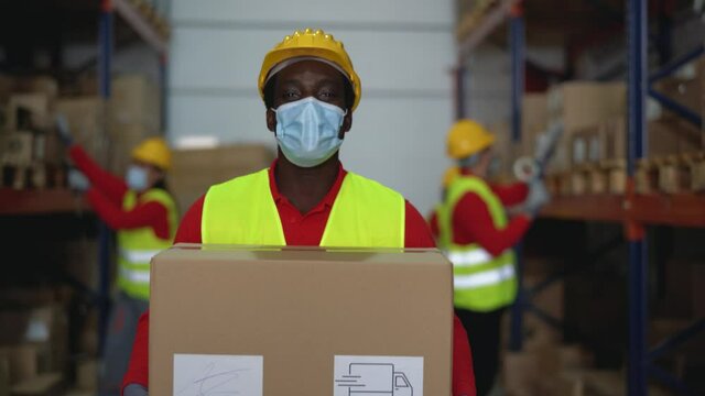 Black man working in warehouse loading delivery boxes while wearing face surgical mask during corona virus pandemic - Logistic and industry concept