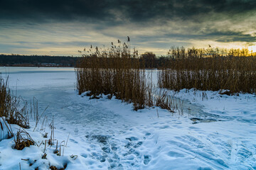 Frozen fishing place in winter at the Pogoria II Lake with heavy clouds and sun over horizon.