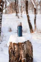 Blue thermos standing on a snow-covered stump. Camping vacuum flask in the winter forest during the day. Vertical photo