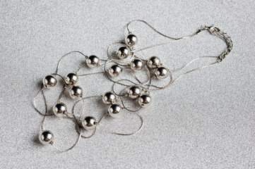 Women's jewelry in the form of silver beads on a silver thread with a clasp, photographed against a silver background.