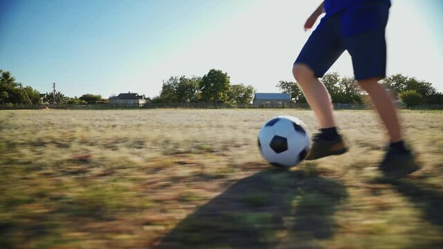 The boy kicks the ball. The kid is training to dribble the ball on a large football field. Football concept. The football player scores the ball into the goal. Street sports in a poor area. Childhood