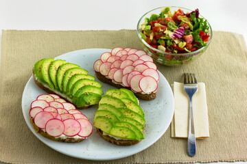 Slices of peasant bread with sliced avocado and radishes on a plate with a salad on the side. 