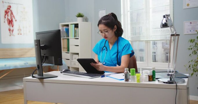 Pretty female nurse using digital tablet, checking documents in on-call room. Adult Asian woman comparing medical reports on gadget and computer while sitting at table in hospital. Technology.