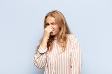 blonde woman feeling disgusted, holding nose to avoid smelling a foul and unpleasant stench