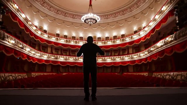 Back view of actor on stage. Man practicing speech with gestures in empty auditorium with red chairs. Performer is reheases in theater.