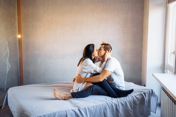 Obraz premium Attractive, young couple of lovers having sex at home in the bed in bright room. Girl kissing her boyfriend oddly, gently pulling back his lower lip. Celebrating Valentine's Day - February 14