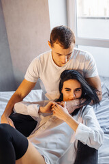 Young heterosexual couple of young lovers indulge in bed at home. Girl lies on legs of boyfriend and hugging his face with hand. Dressed in white shirts and dark pants. Pure enjoyment of each other