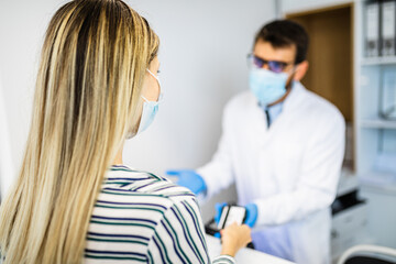 Young male practitioner with face protective mask working at clinic reception desk and talking with patient or visitor.