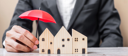 Businessman hand holding red Umbrella cover wooden Home model on table office. Property insurance and real estate concepts