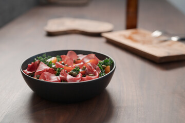 Salad with kale, bresaola and cherry tomatoes decorated with capers in black bowl on walnut wood table