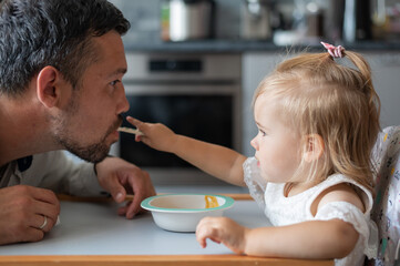 Cute little girl is feeding her dad in the kitchen