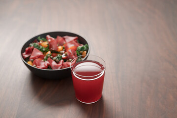 Salad with kale, bresaola and cherry tomatoes decorated with capers in black bowl and glass of red drink on walnut wood table