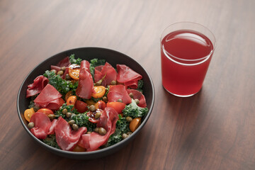 Salad with kale, bresaola and cherry tomatoes decorated with capers in black bowl and glass of red drink on walnut wood table