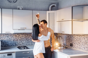 Celebrating Valentine's Day. Loving heterosexual couple with beautiful naked figures and bodies hug, kiss, in the kitchen. Girl holding glass of wine in hands, enjoying the passionate moment