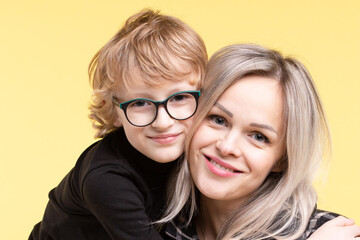 Happy mother with child in glasses hugging, posing on yellow  background. Portrait of smiling Caucasian family mom and son. Blond hair. Horizontal. Copy space. Family, motherhood, vision concept.