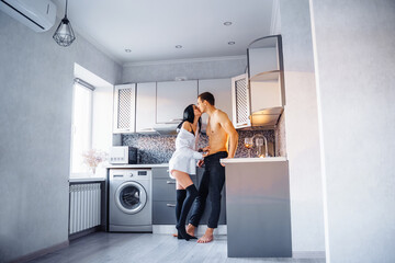 Attractive, passionate, handsome girl and guy having love in the kitchen. Boyfriend with naked muscular torso body gently hugs woman in a white shirt, pressing her against the wall