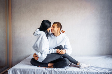 Beautiful young couple in the same white hoodie and black pants lie on the bed hugging. Dreaming of happy future together. Enjoying each other's company. Man gently hugs his beloved from back
