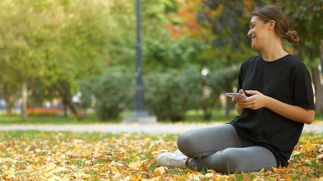 Slow Motion Young beautiful woman sits on lawn in autumn city park, looks and flips through the photos in his smartphone, she smiles, laughs and she is in a good mood