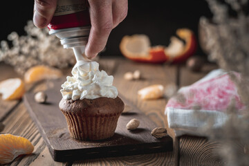 A hand is decorating a appetizing fresh cupcake with whipped cream on a wooden rustic table