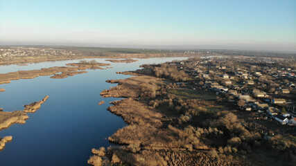 The estuary of a river with blue water. There are dry grass, reeds and trees on the bank and in the middle of the river. There are village with residential houses on the shore.