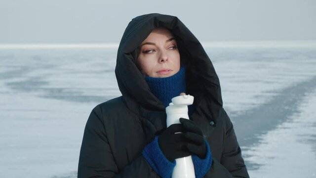 Young woman in warm down jacket with a hood, drinks hot drink from thermos in cold winter on arctic background. Portrait of a female warming herself with hot tea on background of frozen lake or river