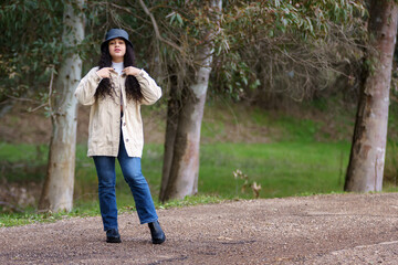 Fototapeta premium Chica adolescente hispana de pelo negro y rizado en el camino en el bosque en invierno con abrigo y sombrero.