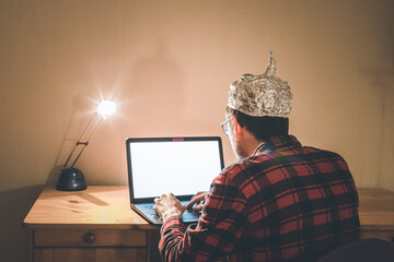 Conspiracy theory concept: young man with aluminum cap searching the internet, sitting lonely in...
