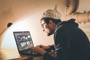 Conspiracy theory concept: young man with aluminum cap searching the internet, sitting lonely in...