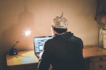 Conspiracy theory concept: young man with aluminum cap searching the internet, sitting lonely in...