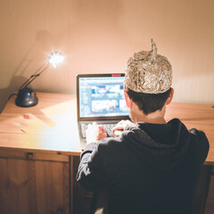 Conspiracy theory concept: young man with aluminum cap searching the internet, sitting lonely in...