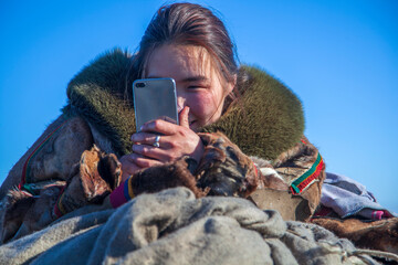 A young girl, in the national winter clothes of the northern inhabitants of the tundra, takes a selfie on a smartphone