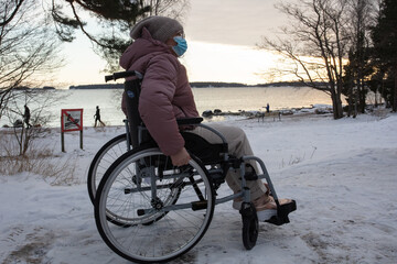 A woman in a wheelchair on a walk by the sea, in winter.
, she is wearing a medical mask. Concept of protection against coronavirus