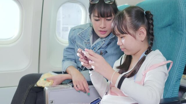 Asia mother talk with her daughter while she is playing game on smartphone at the economy class of airplane. Zoom in to daughter and mom point on phone screen. Passenger travel on the vacation.