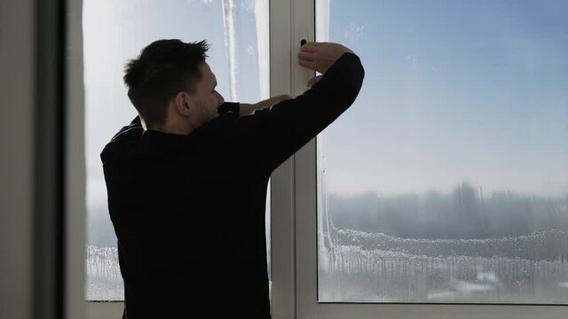 Caucasian man with difficulty opens sliding aluminum sash of balcony window on frosty sunny day, view of city opens, brunette has steam from his mouth because of cold air