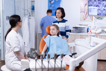 Obraz premium Parent with little girl listening pediatric dentist talking about mouth hygine in dental office. Child with her mother during teeth check up with stomatolog sitting on chair.