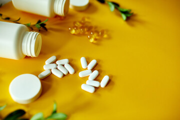 Flat lay of pills, fish oil, vitamins with green leavws on yellow background