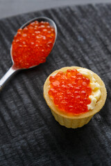 Tartlet with butter and red caviar on a black round board next to a spoon with caviar.