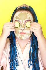 Woman with blue afro braids face in a mask made of green clay with cucumber circles on her eyes on a yellow background.