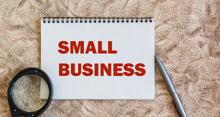 The text SMALL BUSINESS is written on a notebook and a wooden table, next to it lies a magnifying glass and a pen