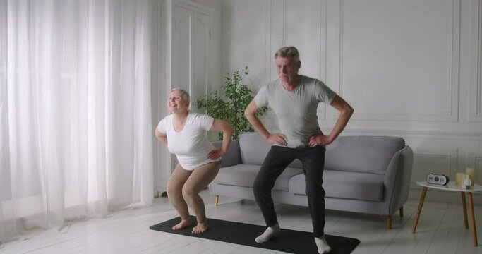 Happy elderly couple doing squats together, enjoying physical activity at home.