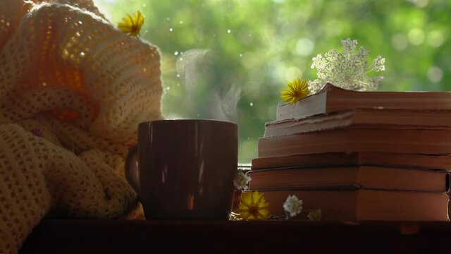 Closeup view 4k video of hot steaming drink standing on windowsill of rural cottage. Near stacked old paper books, some field flowers and yellow cosy blanket