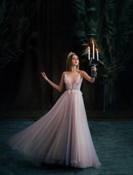 Art photo of medieval girl princess walks runs in dark gothic room. Woman queen is holding candlestick with burning candles in hand. Pink purple dress fly in motion. long blonde hair gold royal crown