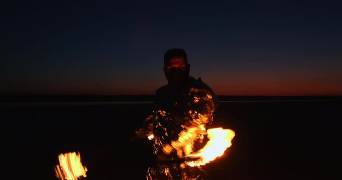 A fiery spinner in a shiny suit twirls fiery torches on chains in front of him, against the backdrop of the sunset sky.
