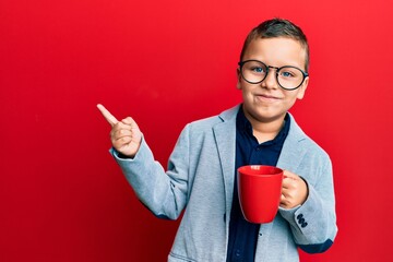 Little kid boy wearing glasses drinking from a red mug smiling happy pointing with hand and finger to the side