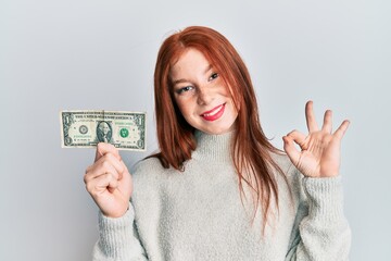 Young red head girl holding 1 dollar banknote doing ok sign with fingers, smiling friendly gesturing excellent symbol