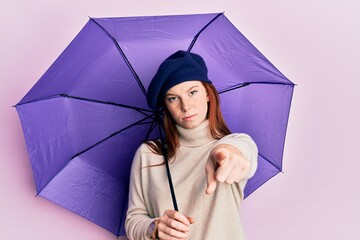 Young red head girl holding purple umbrella wearing fresh beret pointing with finger to the camera and to you, confident gesture looking serious