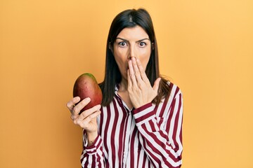 Young hispanic woman holding mango covering mouth with hand, shocked and afraid for mistake....