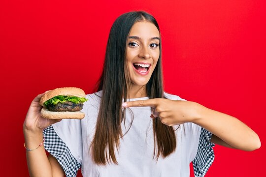 Young hispanic woman eating hamburger smiling happy pointing with hand and finger